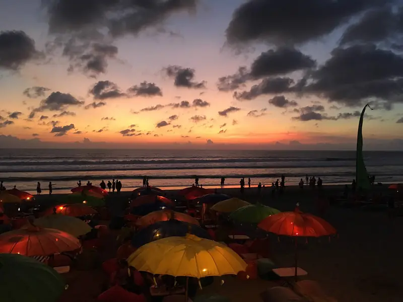 Sunset over Seminyak beach, with rows of coloured parasols and people at the water's edge